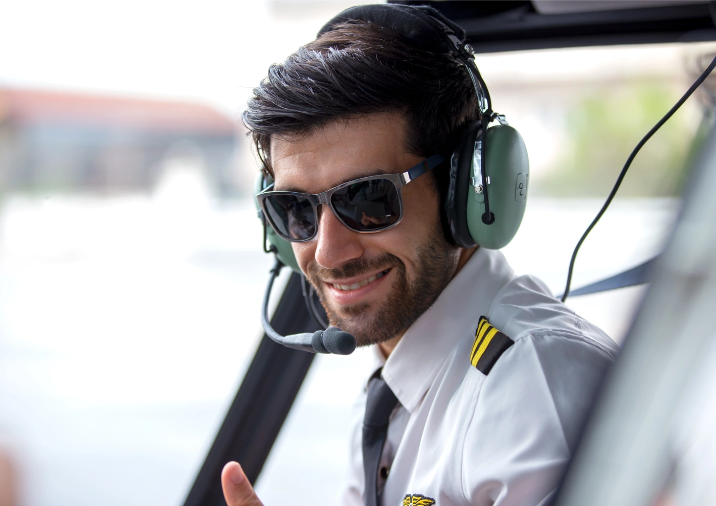 Smiling male pilot wearing headset and sunglasses gives thumbs-up from aircraft cockpit.