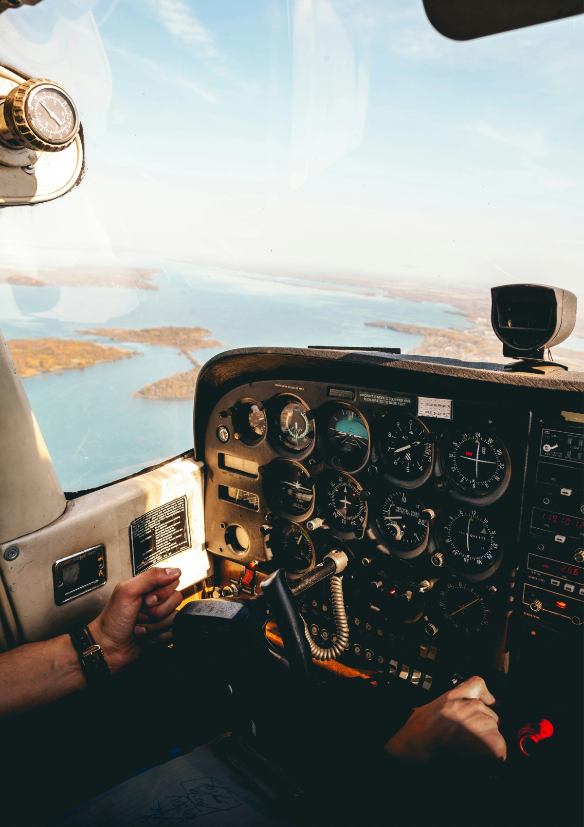 Pilot’s hands on small aircraft cockpit controls overlooking islands and blue water.
